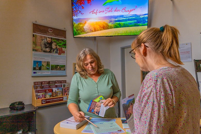 Beratungsgespräch an einem Runden Stehtisch mit Infomaterial vom Geopark. Im Hintergund sind weitere Infoflyer zu sehen und ein Bildschirm mit dem Text: Auf ins Tal der Burgen".