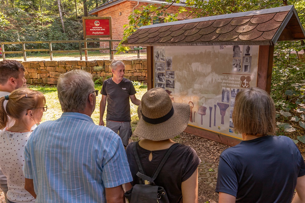 Eine geführte Wandergruppe steht vor einer Infotafel auf der die Werkzeuge des Bergbaus erklärt werden. Im Hintergrund steht das Porphyrhaus.